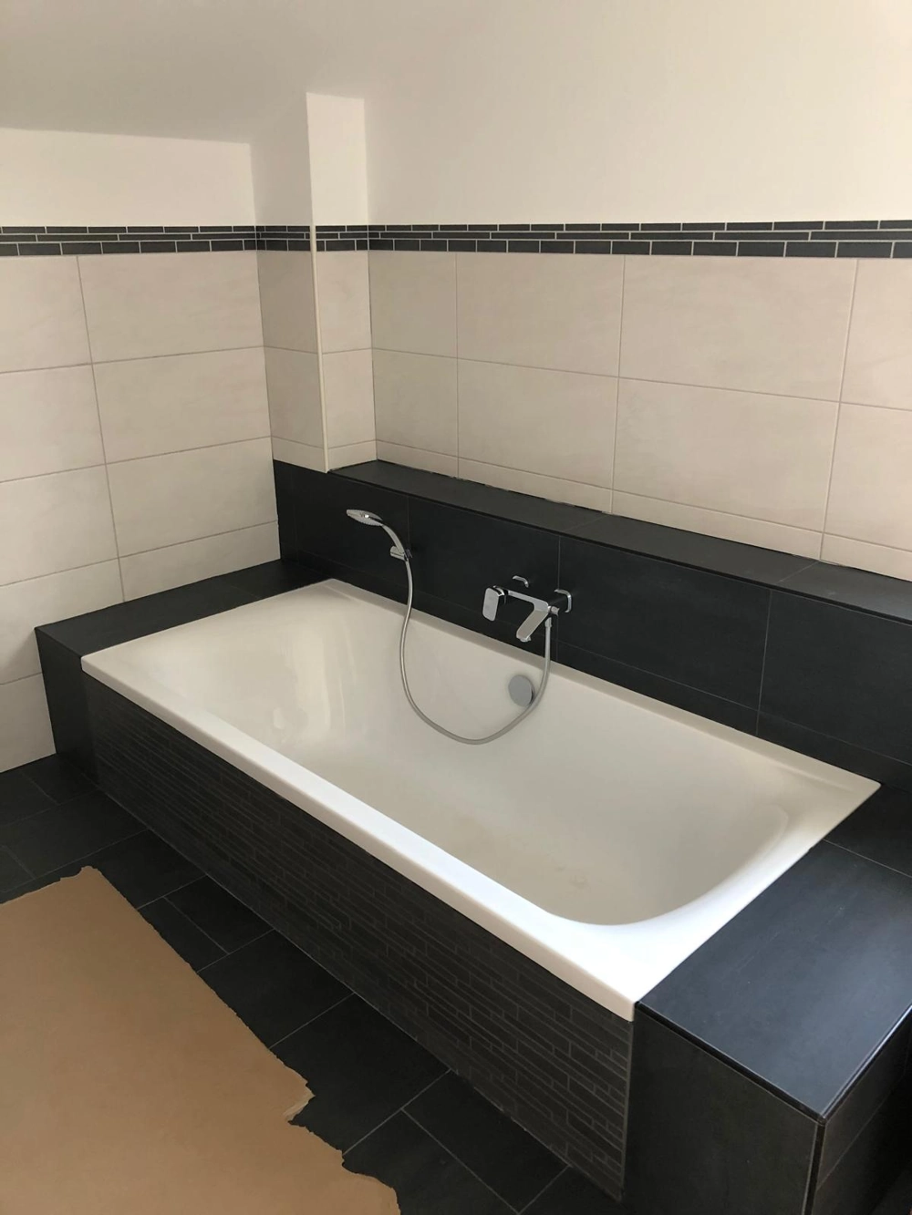 Moderne Badewanne mit schwarzer Fliesenumrandung, Wandarmatur und Handbrause bei Fliesenarbeiten und Sanitärinstallation