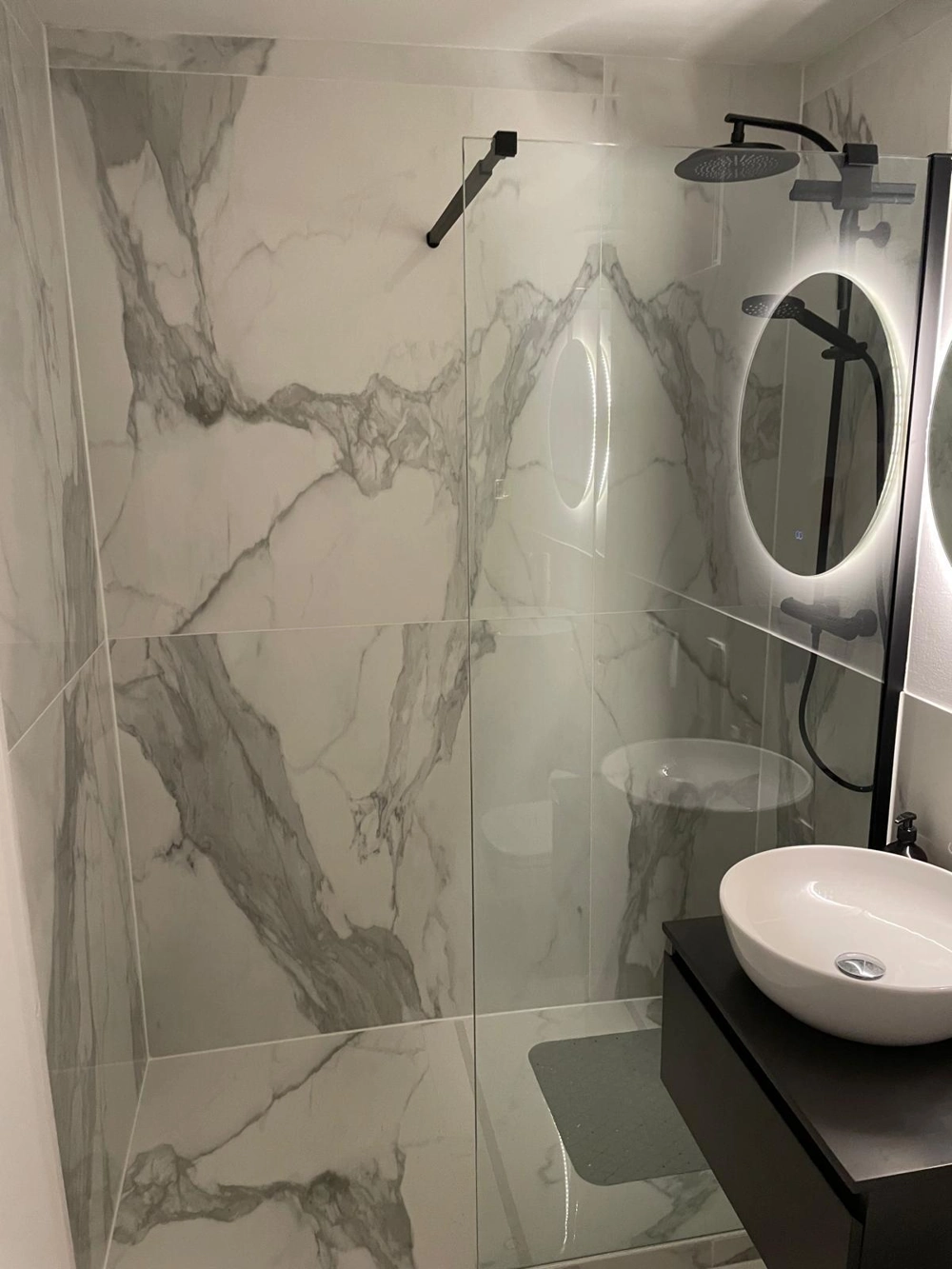 Modernes Bad mit begehbarer Glas-Duschwand, schwarzer Armatur, Rundspiegel mit LED-Beleuchtung und großformatigen Fliesen Fliesenarbeiten