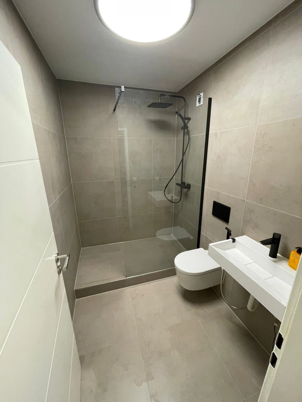 Modernes Badezimmer mit bodengleicher Dusche, rahmenloser Duschkabinen, großformatigen grauen Fliesen, schwarzer Armatur und Wand-WC