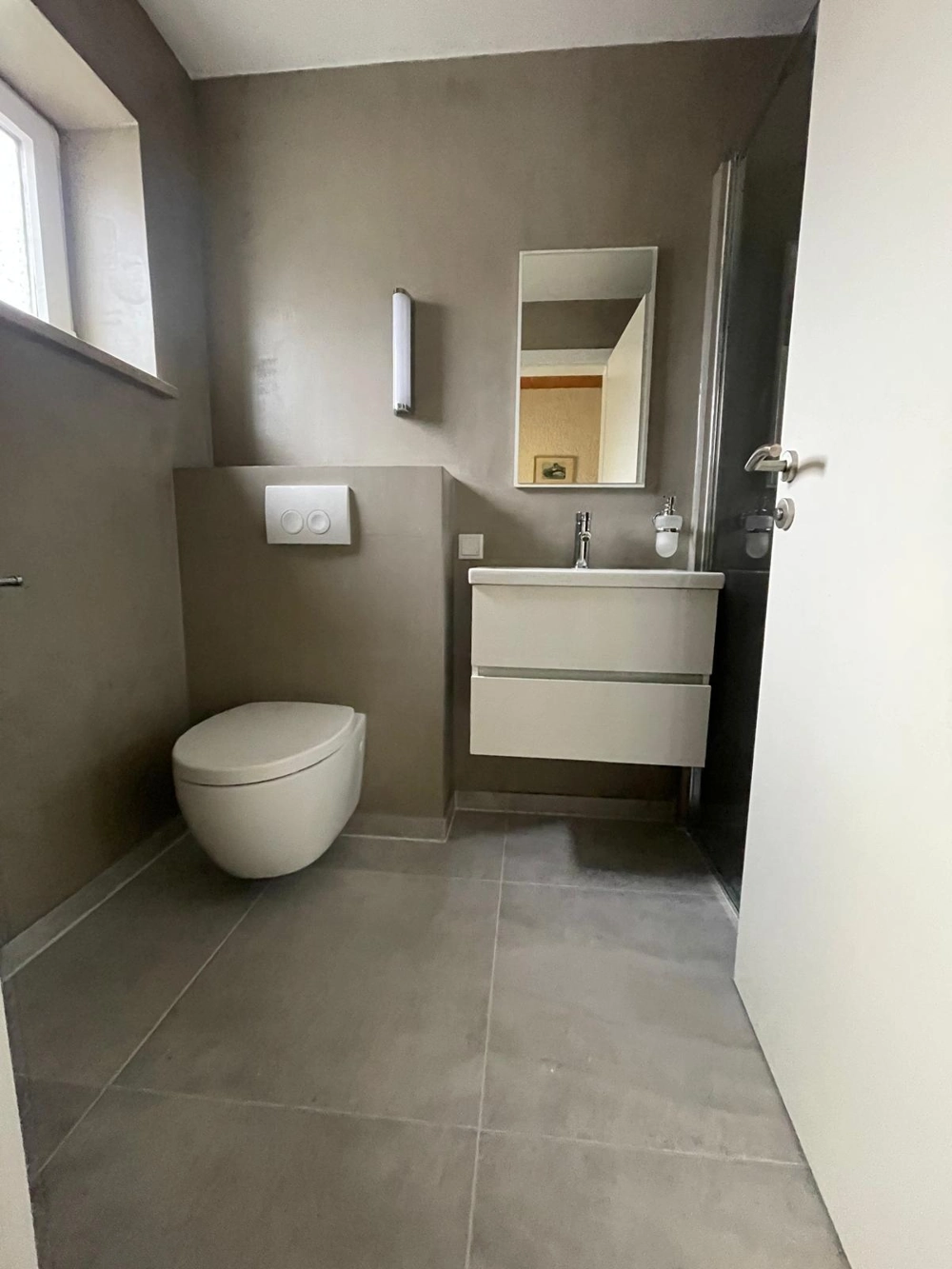 Modernes Bad mit grauen Bodenfliesen, wandhängendem WC, Waschbeckenmöbel, Spiegel und Duschkabine nach Fliesenarbeiten