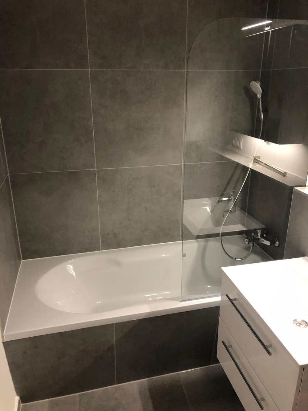 Modernes Badezimmer mit eingelassener Badewanne, Duschwand aus Glas, dunkelgrauen Fliesenarbeiten und Sanitärinstallation